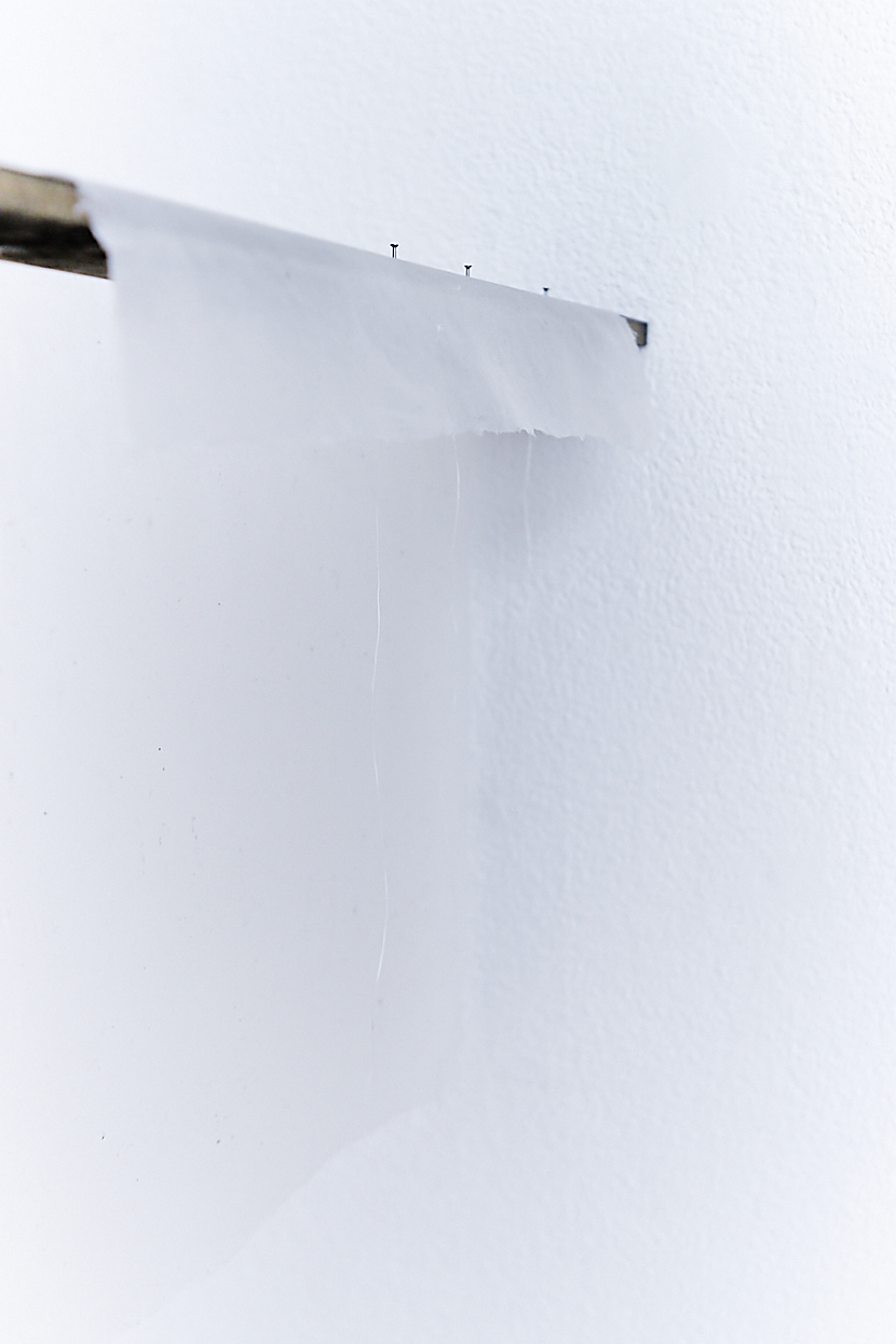 a dark brown wooden board hanging off a white wall. Three steel nails attached on the wood with a white handmade paper