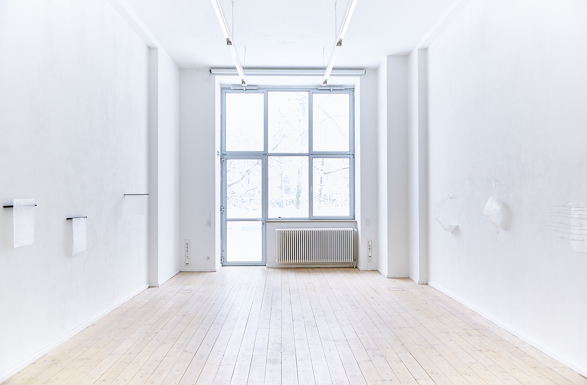 art exhibition installation view of a room with light brown wooded floor and bright window in the center back. On the white walls on the left and right there are six delicate white handmade paper art works with a black wire line structure