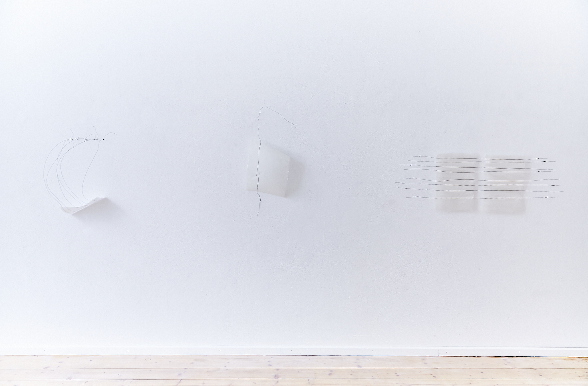 white wall with three delicate white paper artworks on the wall. Some black wire lines are attached to the handmade white paper artworks on the wall. Light brown wooden floor on the bottom of the image