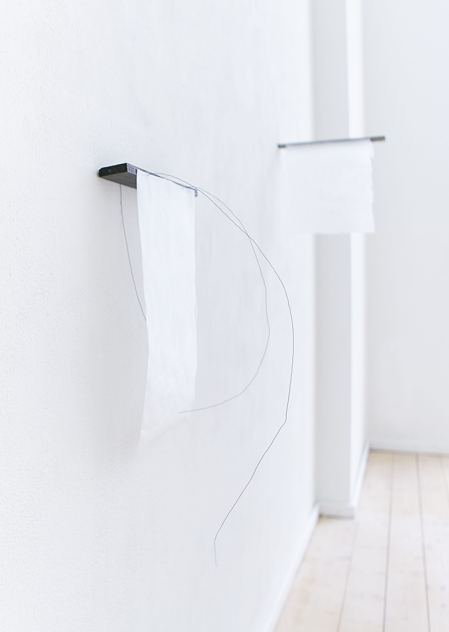 two black pieces of wood on a white wall. Three black thin wire lines grow out of them into a light room which has a wooden light brown floor. Two pieces of transparent handmade papers hang off the wood on the wall