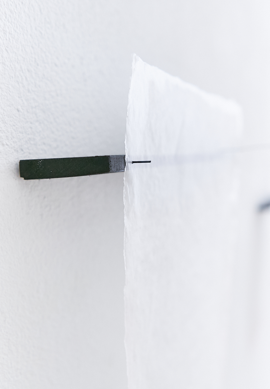 side view of a black piece of wood on the wall. Attached to it a transparent piece of handmade paper which is punched through a small black nail which sits in the black wood